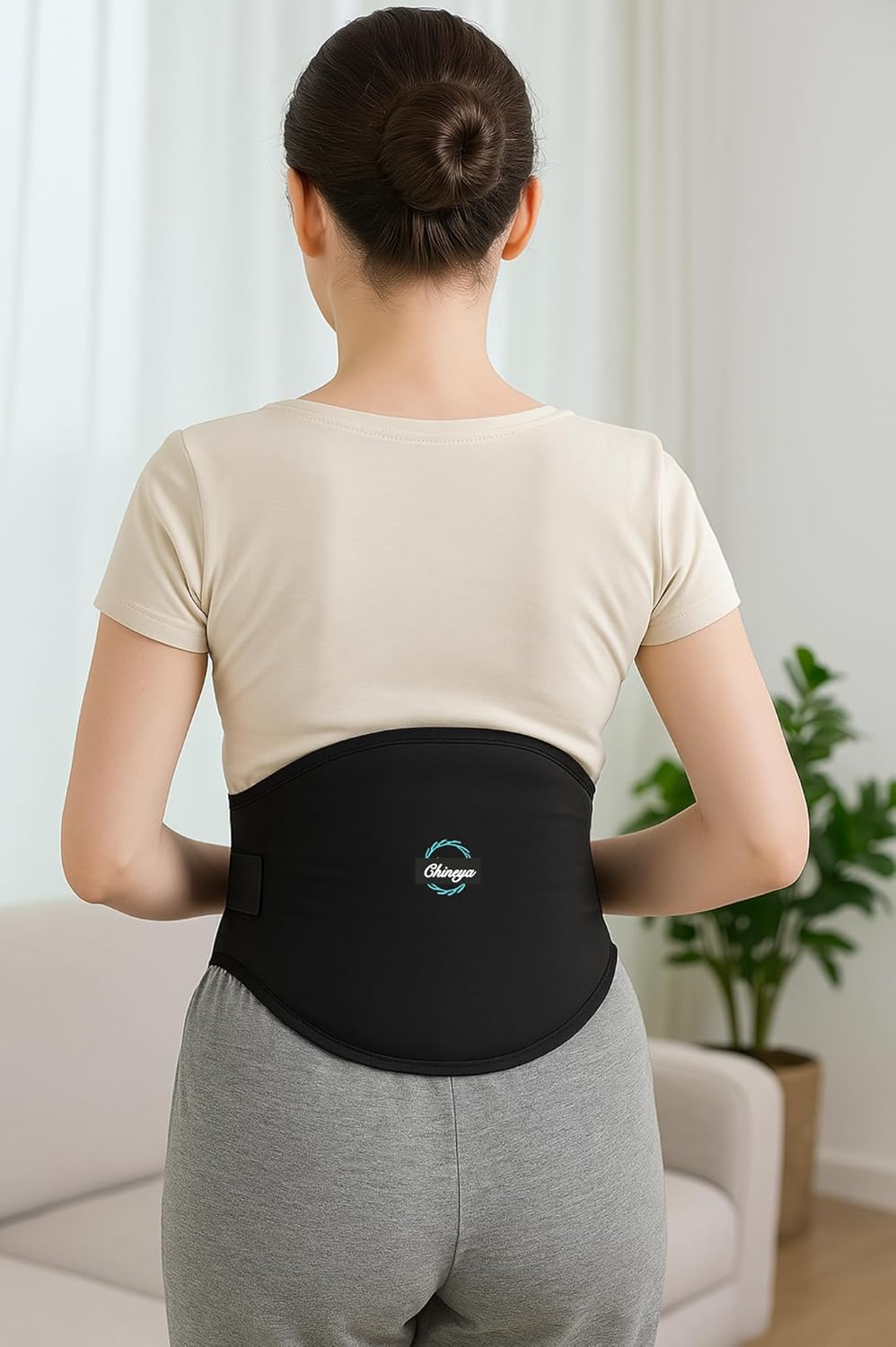 Person wearing a black back brace with a beige shirt and gray pants in a room with a plant and sofa.